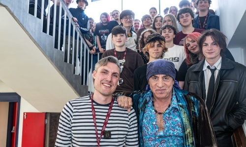 Steve Van Zand stands at the bottom of the stairs in LIPA Sixth Form College with students behind, smiling.